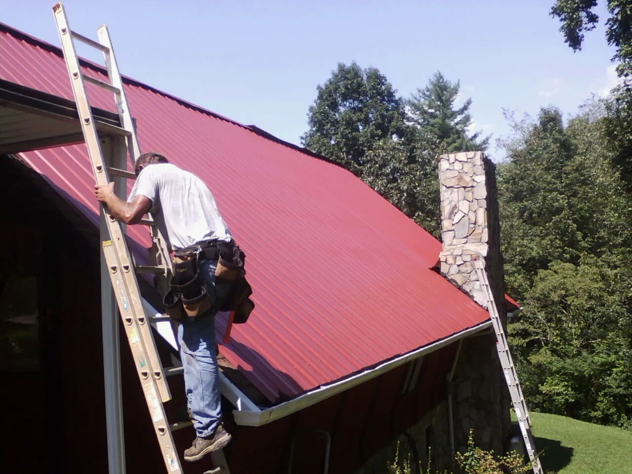 Roofer on a ladder beside a red metal roof and stone chimney — Soffit & Fascia Repair by Roofing Brothers in Natural Bridge Station